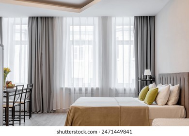 The design of a modern bedroom with a large bed and large panoramic windows. A glass table with yellow flowers in a vase, fruits and croissants. Large chairs. - Powered by Shutterstock