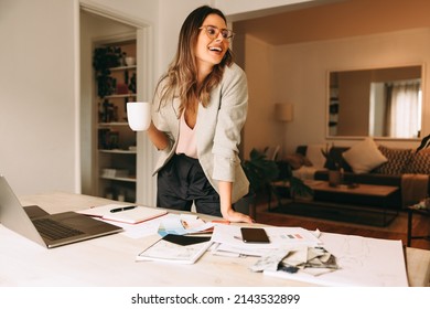 Design professional taking a coffee break in her home office. Creative businesswoman looking away happily while standing behind her desk. Cheerful female designer planning a new project remotely. - Powered by Shutterstock