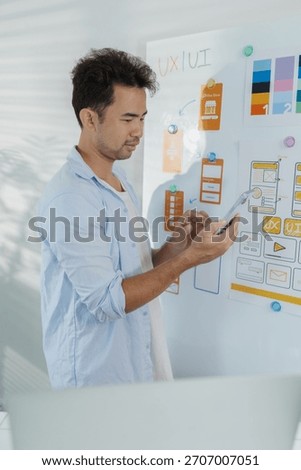 A designer is developing UX UI layouts, reviewing mobile app prototypes, and arranging interface elements on paper and board. He is focused on user experience and digital creativity.