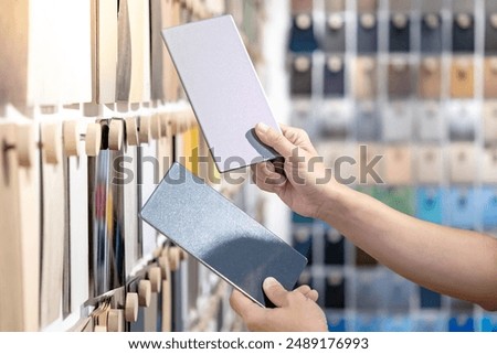 Designer hands compare two gray laminate sheets nearby material swatch wall display in material library. Choosing color board from collection for interior architecture and furniture finishing design