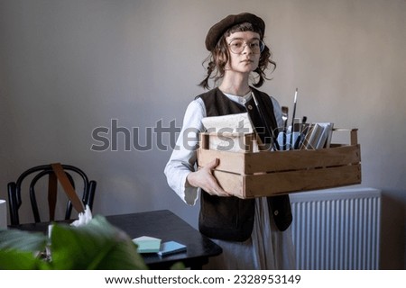 Designer illustrator girl preparing for freelance drawing order looking at camera. Creative artist young woman in stylish beret holds wooden box with art tools, materials, paints for drawing at home. 