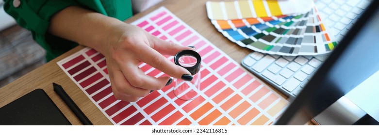 Designer with magnifying glass works with pantone scale in art studio. Computer, drawing tablet, lens and manager woman hands on office desk, top view.