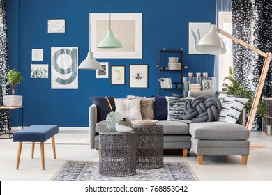 Designer metal tables in living room with navy blue stool near grey corner sofa and lamp