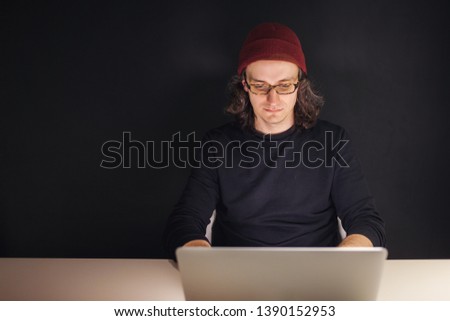 Designer or programmer in the workplace. young man in a hat and glasses sitting at a laptop. dark background