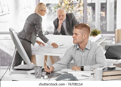 Designer Team At Work In Office, Young Architect Sitting At Desk With Drawing Pad, Older Colleagues Working On Architectural Plan.?