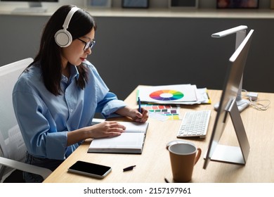 Designer Workplace Concept. Portrait of busy focused Asian woman in glasses and wireless headset writing in notebook sitting at desk table with pc personal computer in modern office, profile side view - Powered by Shutterstock