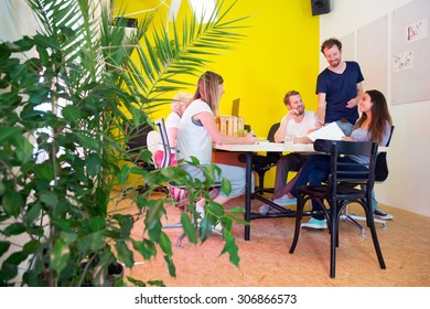 Designers, Sitting In At A Large Table In A Creative Environment And Office, Surrounded By Tack Boards With Drawings, Plants And A Bright Yellow Wall