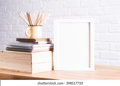 Desk With Blank White Picture Frame And Pencils In Iron Mug Placed On Books And Wooden Box. Mock Up