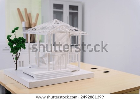 Desk of an engineer displays a concrete house structure under construction. A toy house model sits nearby, surrounded by blueprints and tools, depicting the foundation, roof, and structural elements.