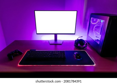 Desk with gaming setup. Display with isolated screen for mockup. Gaming PC, headset, keyboard, mouse and joypad on desk. Purple led light on wall
