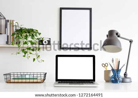 Desk with laptop computer, poster frame, supplies, notebooks, ivy on a shelf and white wall for mock up or copy space.