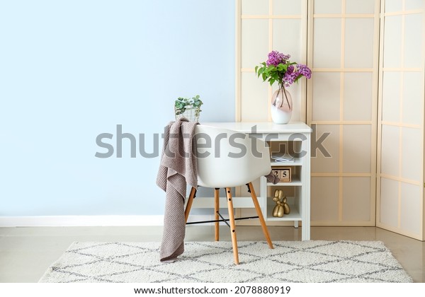 Desk with lilac flowers in vase and folding screen\
near blue wall
