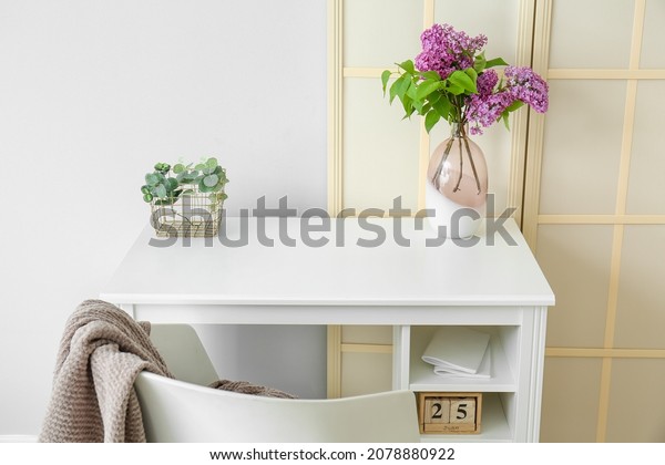 Desk with lilac flowers in vase and folding screen\
near light wall
