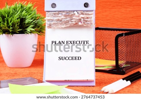 A desk notepad features PLAN EXECUTE SUCCEED on an orange background. It sits next to a green plant, a mesh holder with notes, and pencils.