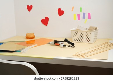 Desk set for a creative DIY project with a hot glue gun, colorful paper, and sticks. Red hearts on the wall suggest a love or Valentine's theme. Bright, clean composition perfect for hobby concepts.