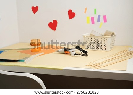 Desk set for a creative DIY project with a hot glue gun, colorful paper, and sticks. Red hearts on the wall suggest a love or Valentine's theme. Bright, clean composition perfect for hobby concepts.
