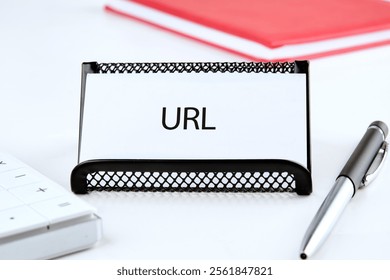 A desk setup featuring a black wire note holder with a card labeled 'URL.' Nearby are a keyboard and a silver pen, illustrating a tidy and organized workspace for digital tasks.