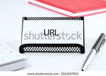 A desk setup featuring a black wire note holder with a card labeled 'URL.' Nearby are a keyboard and a silver pen, illustrating a tidy and organized workspace for digital tasks.