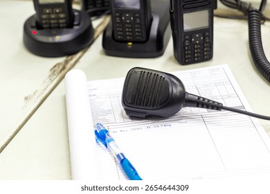 A desk setup featuring professional two-way radios, a detached microphone, and a logbook with a pen, indicating essential communication and record-keeping for operations or security.