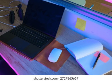 Desk setup with a laptop, mouse, notebook, and pen illuminated by colorful neon lights