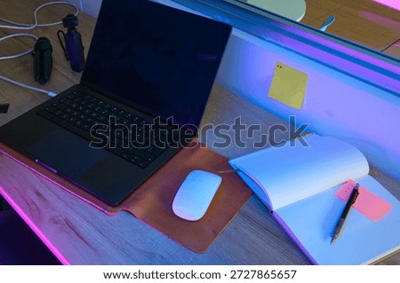 Desk setup with a laptop, mouse, notebook, and pen illuminated by colorful neon lights