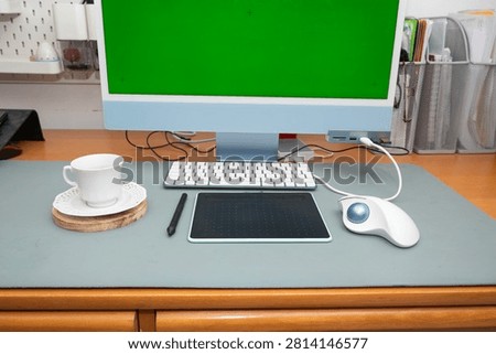 Desk view with a modern computer featuring a customizable green screen. A graphic tablet with stylus. A keyboard. A trackball mouse. And a coffee cup