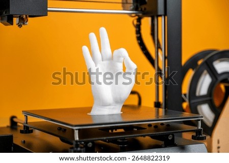 Desktop 3D printer printing raised human hand sculpture using white filament, concept of digital fabrication, prototyping, futuristic manufacturing technology inside modern studio with vibrant yellow 