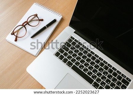 desktop top view, laptop with black display, notepad, pen, glasses, paper clip, bank card, phone on a wooden table