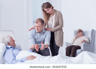 Desolate Grandson Talking To His Grandfather Who Is Lying In A Hospital Bed, With Other Family Members In The Background