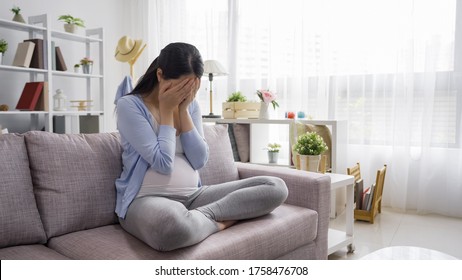 Despair Pregnant Asian Chinese Girl Covering Face With Two Hands While Sitting And Resting On Couch In Bright Cozy Living Room At Home Alone. Maternity Lady Crying In Sofa Feeling Depressed In House