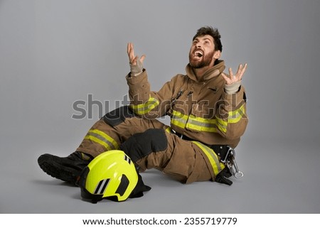 Despaired bearded fireman raising hands, crying after fighting fire in studio. Side view of sooty, emotional, sad firefighter screaming, while sitting on floor, on gray background.Concept of emotions.