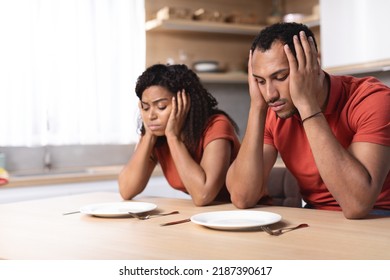 Despaired Sad Pensive Young African American Man And Woman Hold Head With Hands Look At Empty Plates At Table In Kitchen Interior. Diet, Food Crisis And Poverty. Hungry Couple Waiting For Dinner