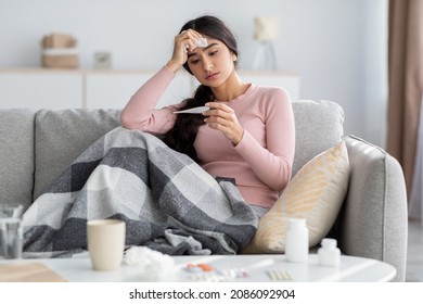 Despaired sad young hindu lady with plaid, suffer from flu, sits on sofa and looks at thermometer in living room interior. Cold, fever, disease treatment at home and covid-19 lockdown, free space
