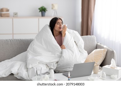 Despaired Sad Young Indian Lady Wrapped In Blanket Sneezes In Napkin, Studies And Works At Home In Living Room Interior With Laptop. Covid-19 Virus, Cold And Flu, Ill And Treated During Quarantine