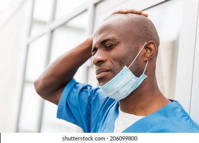 Despaired Surgeon. Depressed Young African Doctor In Blue Uniform Touching Head With Hand And Keeping Eyes Closed While Leaning At The Glass Door