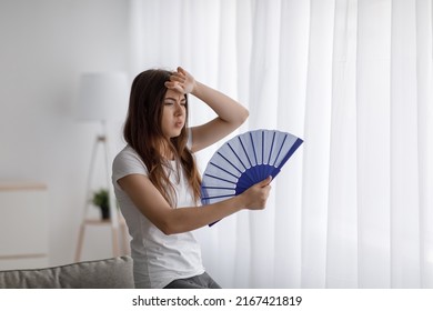 Despaired Tired Millennial Caucasian Lady Suffering From Heat Waves Blowing Fan At Herself, Wipes Sweat With Hand In Room Interior On Window Background. Too Hot At Summer Weather, Overheating At Home