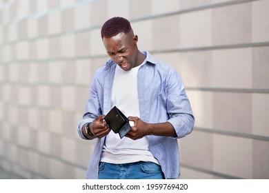 Desperate African American guy looking at his empty wallet near brick wall on city street. Unhappy black man with no cash in his purse outdoors. Economic crisis and bankruptcy concept