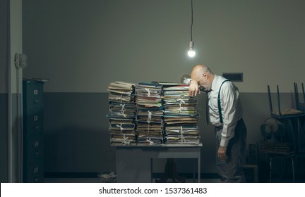 Desperate businessman with lots of paperwork in his messy office at night, he is leaning on a pile of files, bureaucracy and deadlines concept