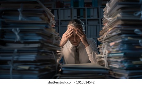 Desperate businessman working in the office late at night and overloaded with work, his desktop is covered with paperwork: business management and deadlines concept - Shutterstock ID 1018514542