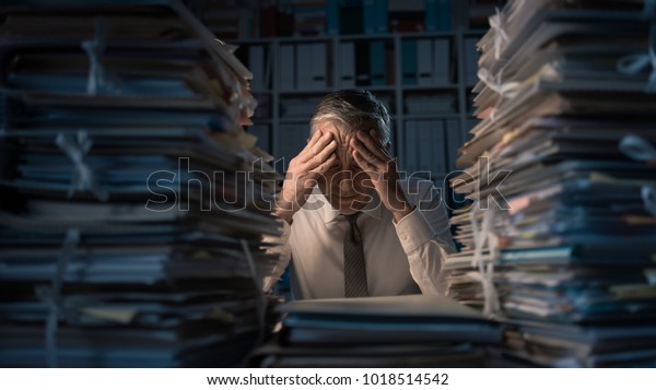 Desperate businessman working in\
the office late at night and overloaded with work, his desktop is\
covered with paperwork: business management and deadlines\
concept