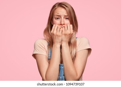 Desperate Caucasian female with worried expression, bites finger nails nervously, finds out tragic news, poses against pink background. Intelligent student worries before answering on final exam