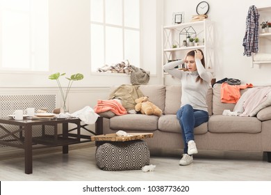 Desperate helpless woman sitting on sofa in messy living room. Young girl surrounded by many stack of clothes. Disorder and mess at home, copy space - Shutterstock ID 1038773620