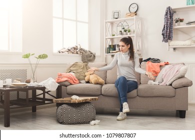 Desperate Helpless Woman Sitting On Sofa In Messy Living Room. Young Girl Surrounded By Many Stack Of Clothes. Disorder And Mess At Home, Copy Space