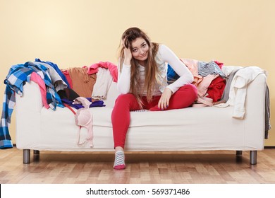 Desperate Helpless Woman Sitting On Sofa Couch In Messy Living Room With Hand On Head. Young Girl Surrounded By Many Stack Of Clothes. Disorder And Mess At Home.