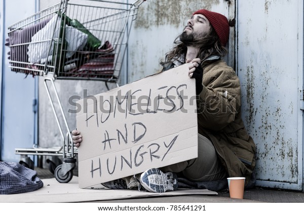 Desperate\
homeless and hungry tramp with a sign sitting on the street next to\
a trolley and begging for food and\
money