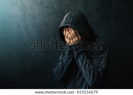 Desperate man in hooded jacket is crying, hands are covering face and tears in the eyes, light of hope shining from his right side