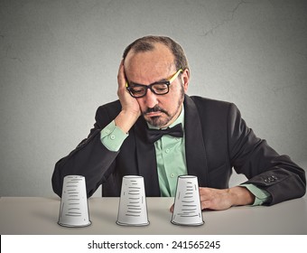 Desperate Sad Middle Aged Business Man With Glasses Sitting At Desk Looking Down Playing A Conjuring Trick Game Isolated Grey Wall Office Background. Human Face Expression. Gambling Risk Concept 