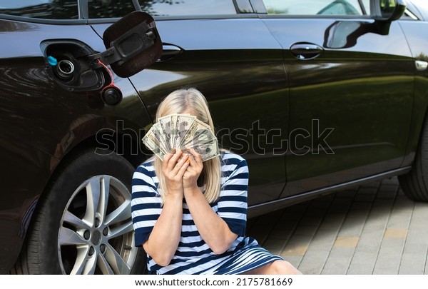 Desperate woman hides face behind cash\
near an open car fuel tank, concept of rising fuel\
prices