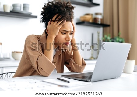 Desperate worried sad african american young woman, using laptop for work or study from home, sitting in kitchen, frustrated looking at screen, failed project, got financial loss, job downgrade