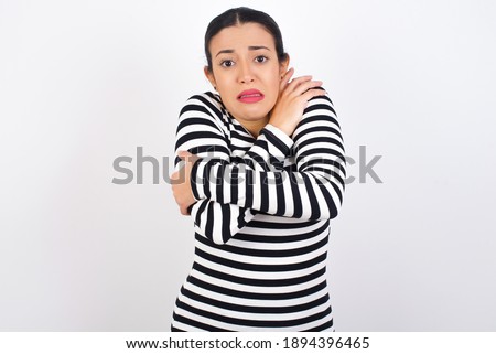 Desperate Young beautiful woman wearing stripped t-shirt against white background trembles and feels cold, hugs oneself to warm up or feels scared notices something terrifying.